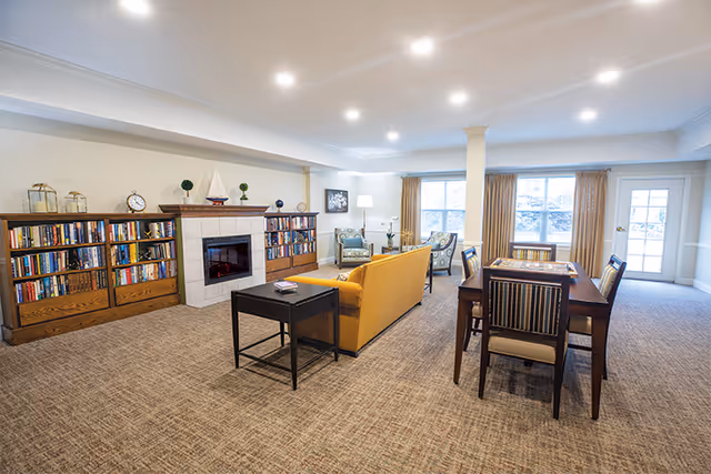 Spacious furnished common room with a yellow sofa, fireplace flanked by bookshelves, seating areas and a dining table under recessed lighting.