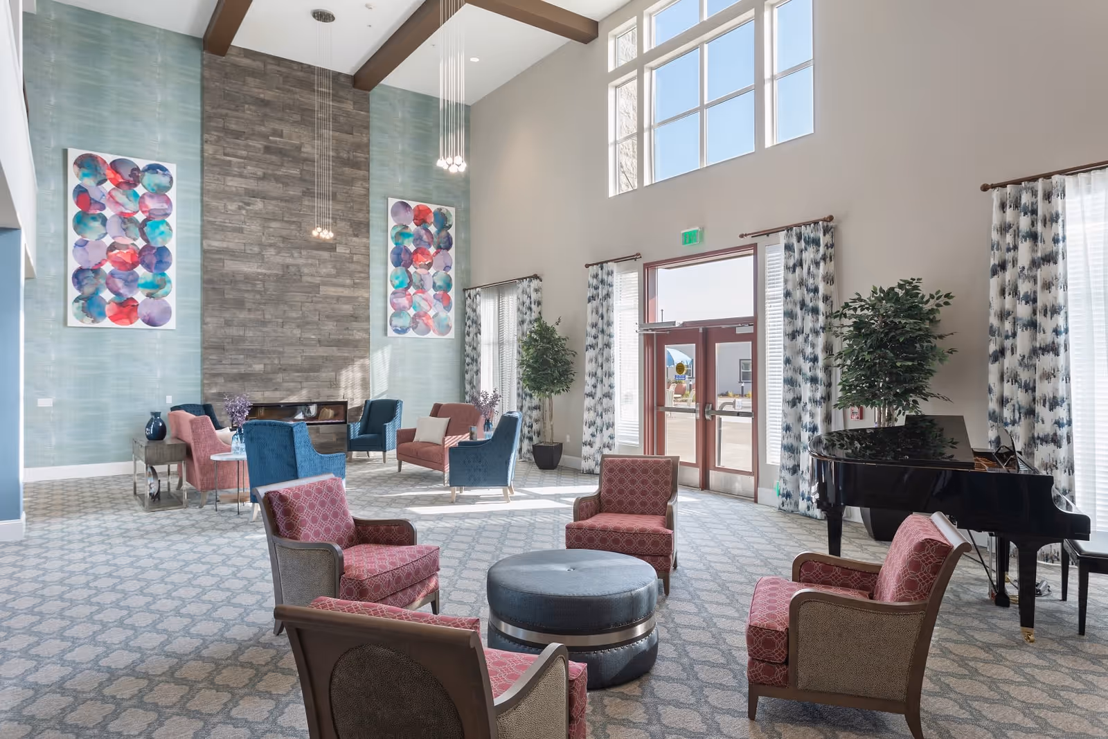 A spacious and well-lit living room area with high ceilings and large windows. The room features multiple seating arrangements including red and blue upholstered chairs, a round ottoman in the center, and a black grand piano near the windows. The walls have decorative artwork and a textured accent wall with hanging pendant lights. Patterned curtains frame the windows and door leading outside.