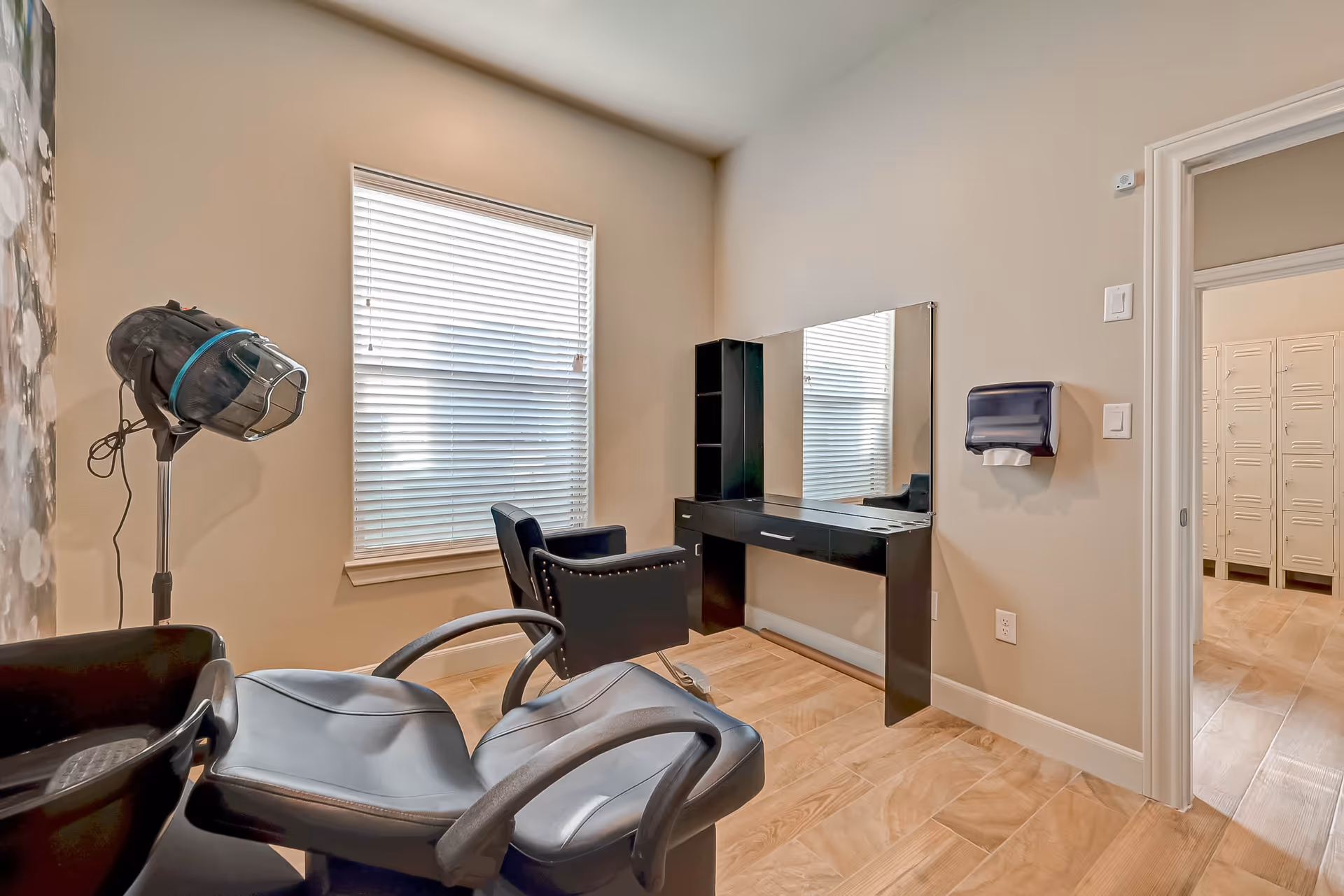 Small salon-style room with styling chairs, a hooded hair dryer, a wall-mounted mirror and counter next to a window with blinds.