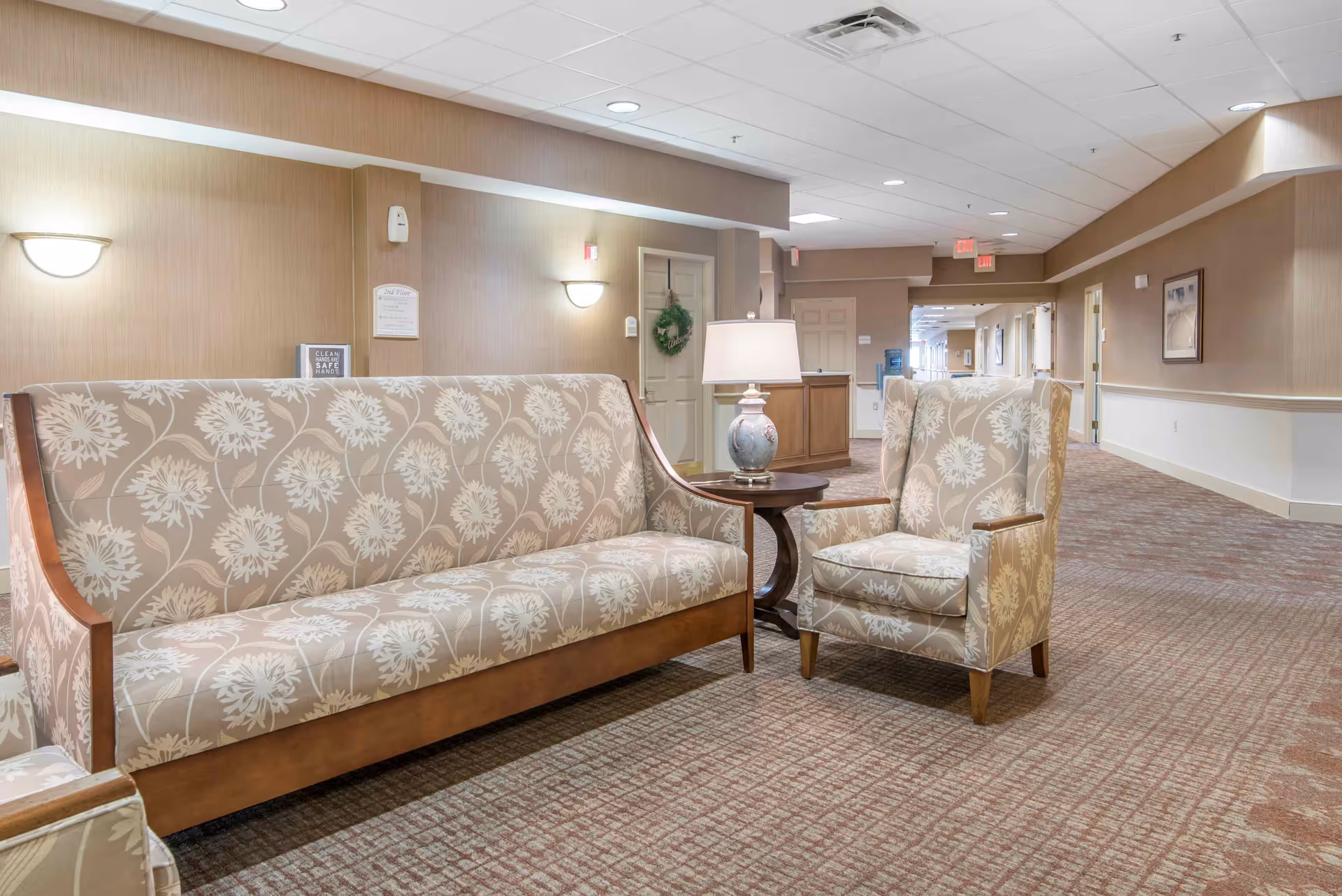 A well-lit hallway in a senior living facility with patterned beige and white upholstered sofa and armchair, a small round wooden table with a decorative lamp, beige walls with wall sconces, and carpeted floor. The hallway extends into the distance with doors and framed pictures on the walls.