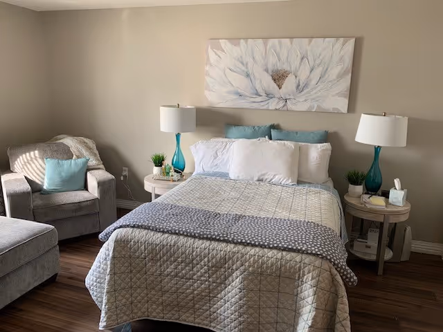 A cozy bedroom with a neatly made bed featuring a quilted bedspread and multiple pillows. On either side of the bed are round wooden nightstands with matching blue glass lamps, small plants, and other decorative items. To the left of the bed is a comfortable armchair with a blue cushion and a matching ottoman. Above the bed hangs a large floral painting with white petals and a brown center. The room has wooden flooring and beige walls.