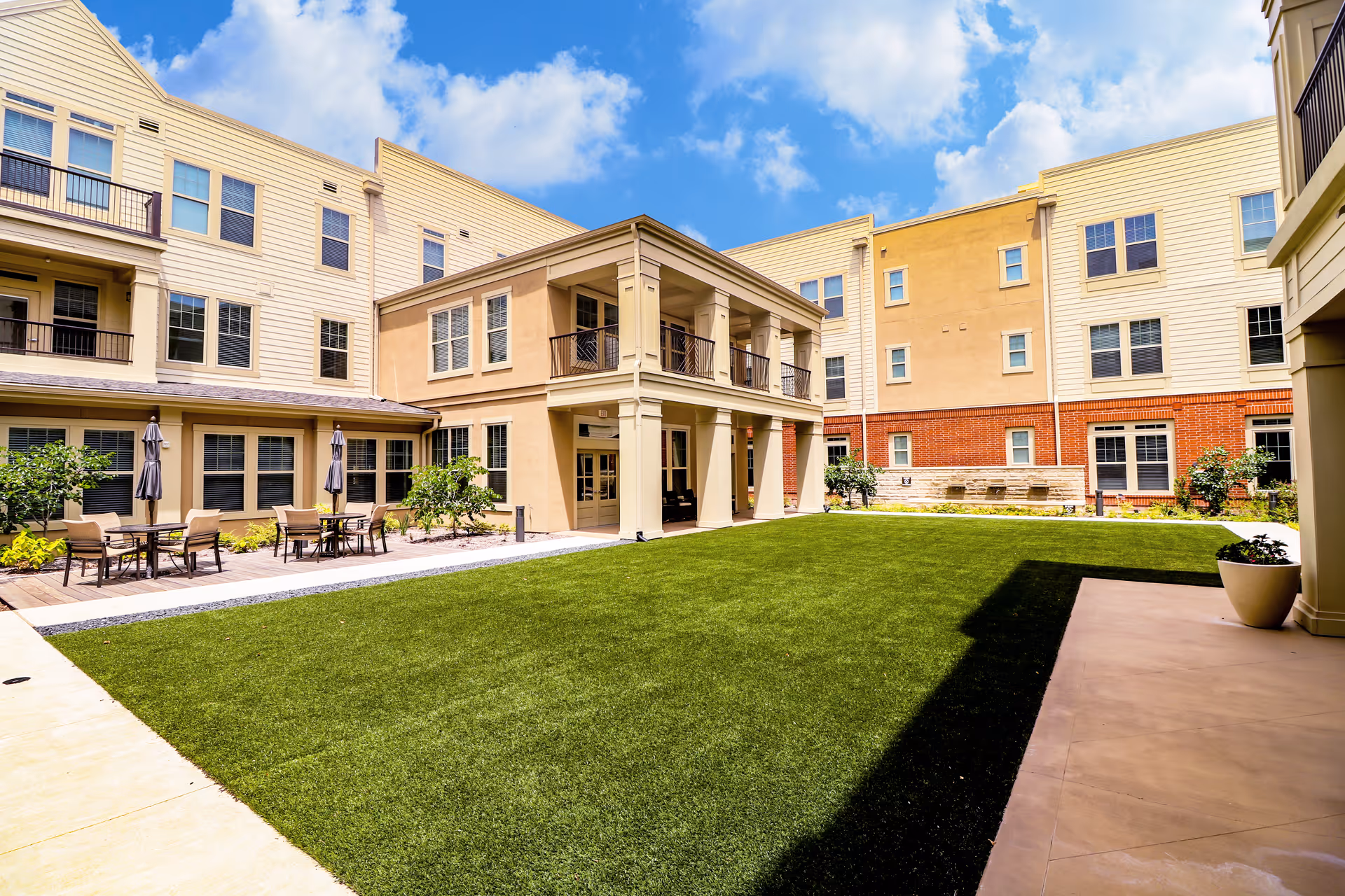 Outdoor courtyard area of an assisted living facility with a well-maintained green lawn, patio tables with umbrellas and chairs, surrounded by a multi-story building under a partly cloudy blue sky.