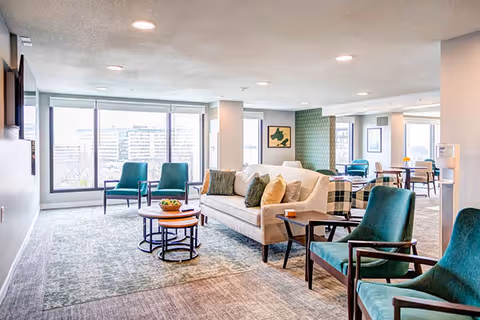 Bright communal lounge with a sofa, teal armchairs, nesting coffee tables, and large windows overlooking the city.