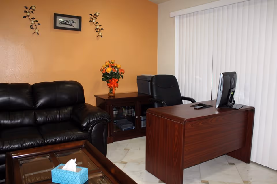 An office area with a dark leather couch, a wooden desk with a computer monitor and a smartphone on it, a black office chair, a wooden cabinet with a printer and a vase of orange and yellow flowers, and vertical blinds covering a window.