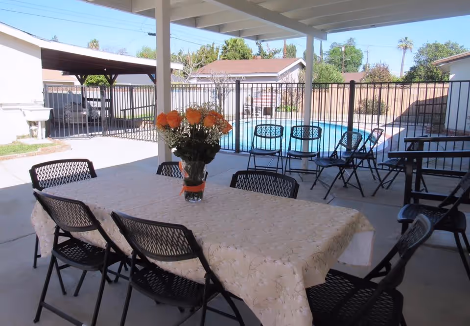 Covered outdoor patio with a long table and chairs, a vase of orange flowers on the table, and a fenced swimming pool in the background.