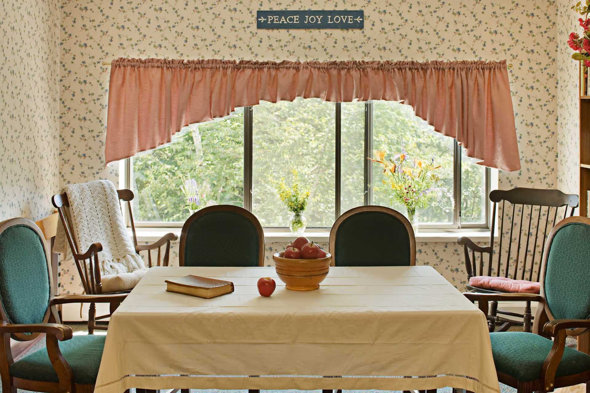 Cozy dining room with a cloth-covered table holding a bowl of apples and a book, surrounded by green-upholstered chairs and rocking chairs in front of a window with a pink valance and floral wallpaper.