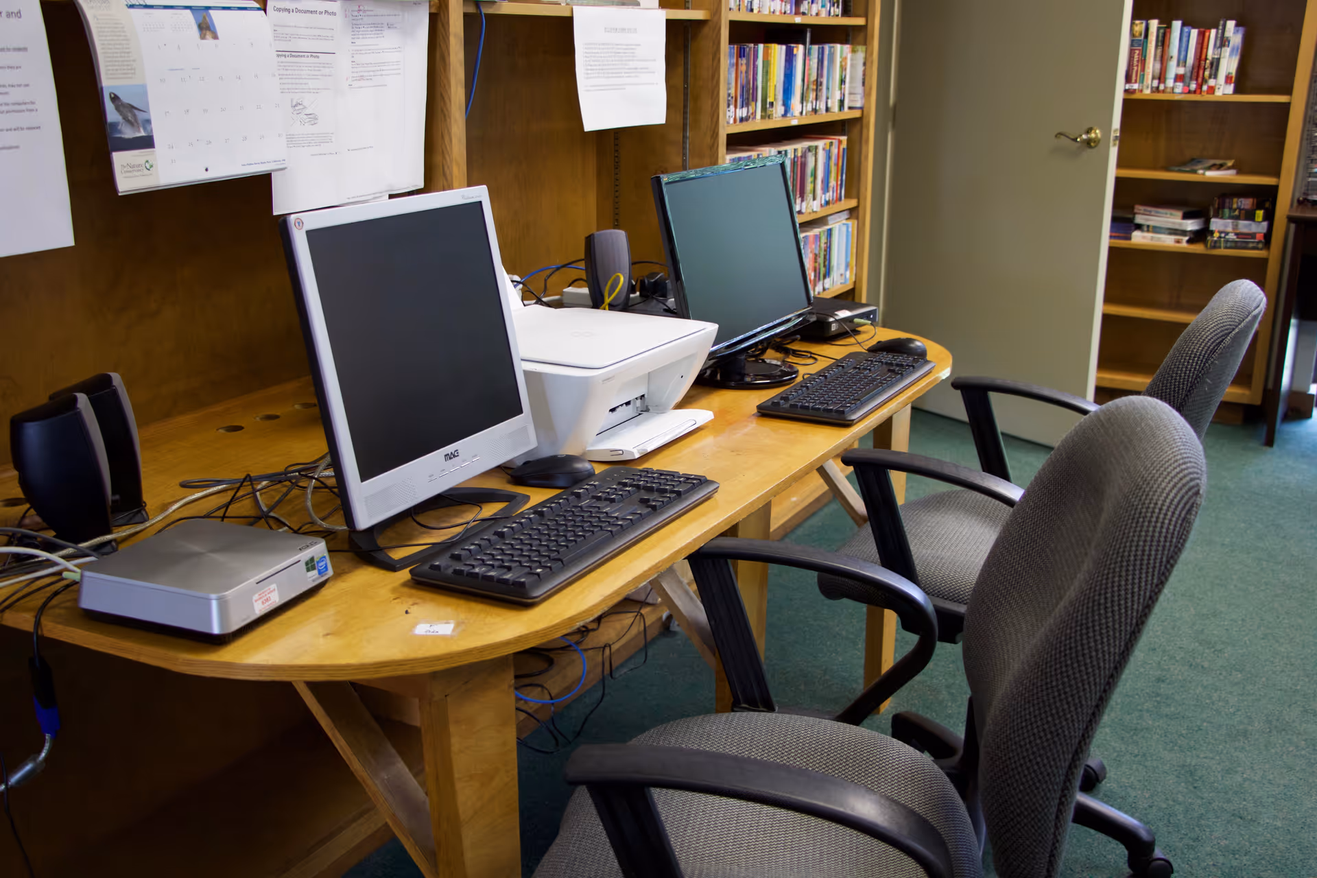 Two desktop computers, a printer, keyboards and office chairs on a wooden desk in a small library or common room.