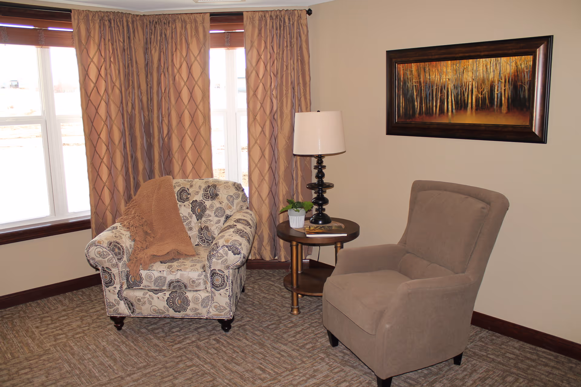 A cozy sitting area with two armchairs, one patterned with floral designs and a brown throw blanket, and the other solid beige. Between them is a round wooden side table with a black lamp, a small potted plant, and a book. Behind the chairs are large windows with beige curtains, and a framed painting of a forest hangs on the beige wall.