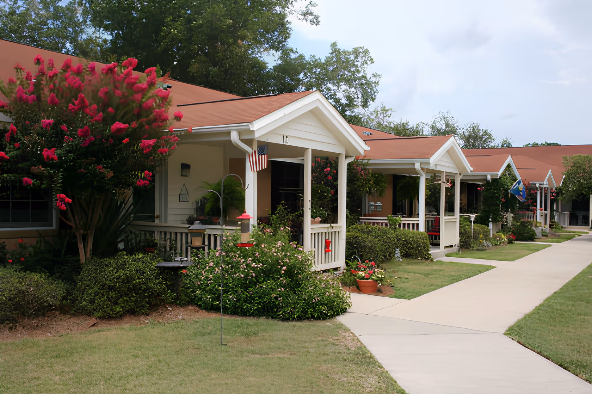 Row of single-story residential units with small covered porches, surrounded by well-maintained gardens and flowering bushes, with a paved walkway in front under a partly cloudy sky.