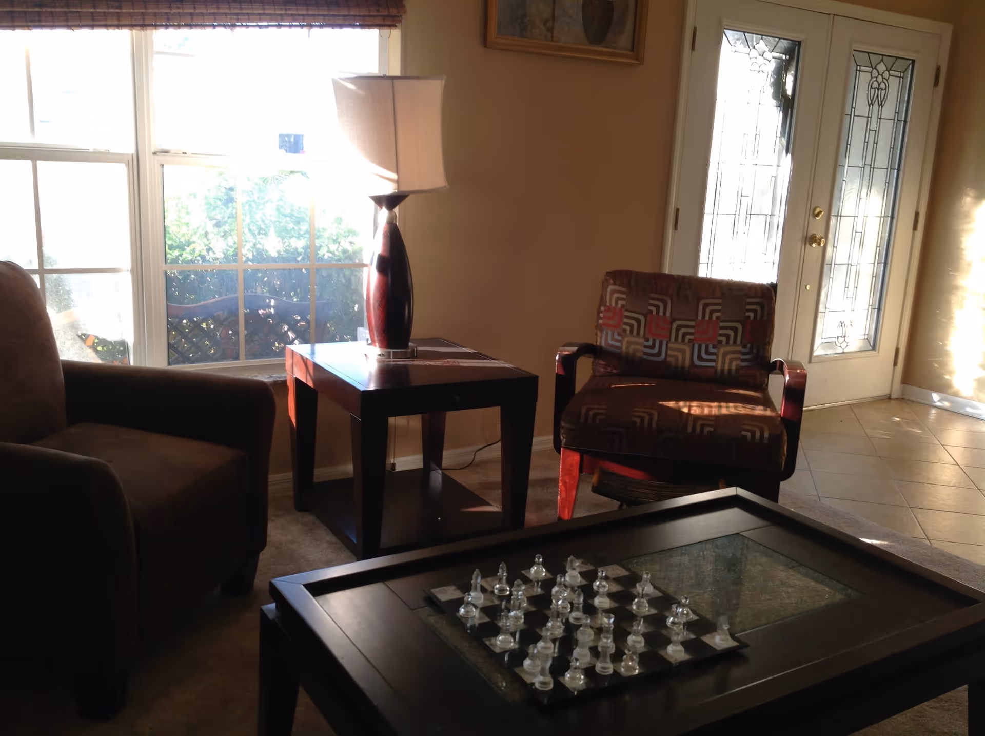 A cozy living room area with two armchairs, a side table with a lamp, a glass chess set on a coffee table, large windows letting in natural light, and a decorative glass-paneled door.