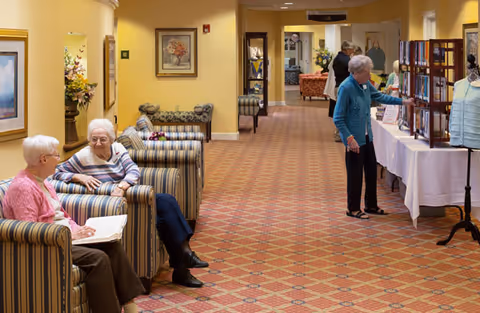 A hallway in a senior living facility with patterned carpet and yellow walls. Two elderly women are seated on striped armchairs on the left side, engaged in conversation. On the right side, another elderly woman is looking at items displayed on tables covered with white tablecloths. The hallway leads to a common area with more seating and floral arrangements.