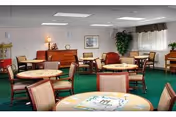 Common area with round game tables and chairs, a piano, and a potted plant in a senior living facility.