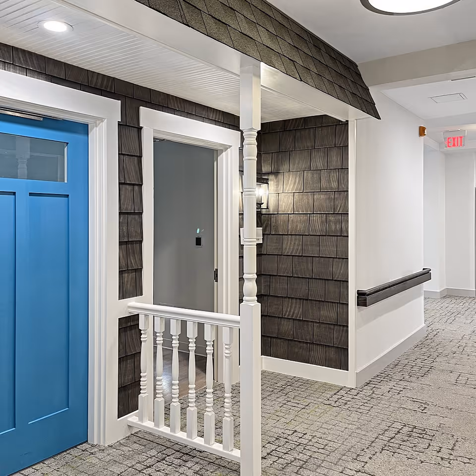Interior hallway featuring a blue door next to a small decorative porch-like facade with shingle siding, white railing, and a wall lantern.