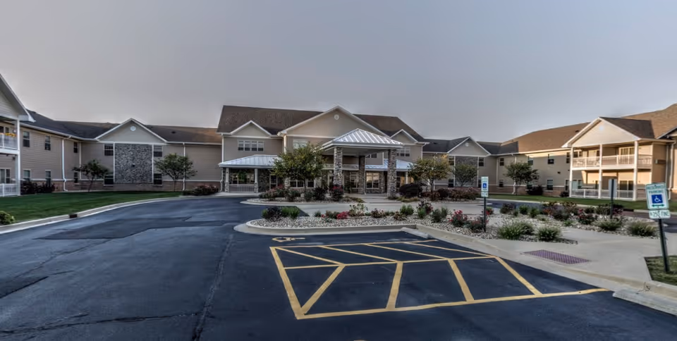 Front exterior view of Lincolnshire Place of Decatur, showing a large two-story building with beige siding and stone accents. There is a covered entrance with stone pillars, landscaped flower beds, and a parking lot with marked handicap parking spaces in the foreground.