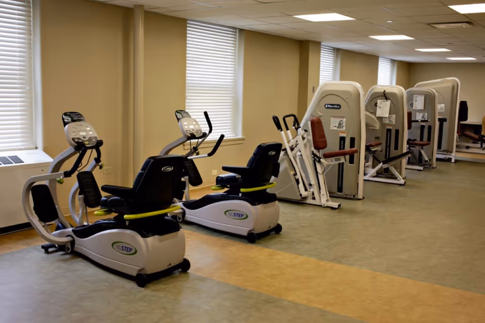 Interior view of a fitness room with exercise equipment including two recumbent bikes and several strength training machines lined up against the wall with windows covered by blinds.