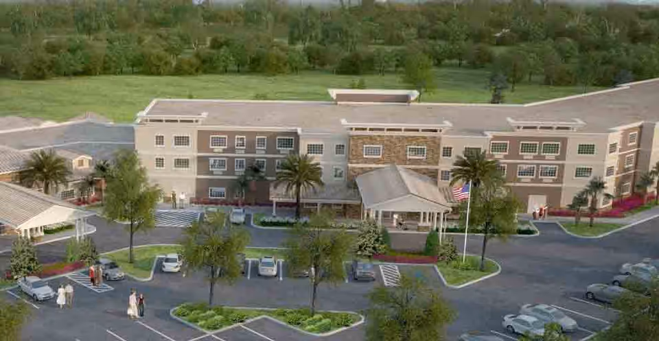 Aerial view of Discovery Village At Stuart, showing a large three-story building with a covered entrance, surrounded by parking spaces, palm trees, and landscaped greenery. Several people are walking near the entrance and parking lot.