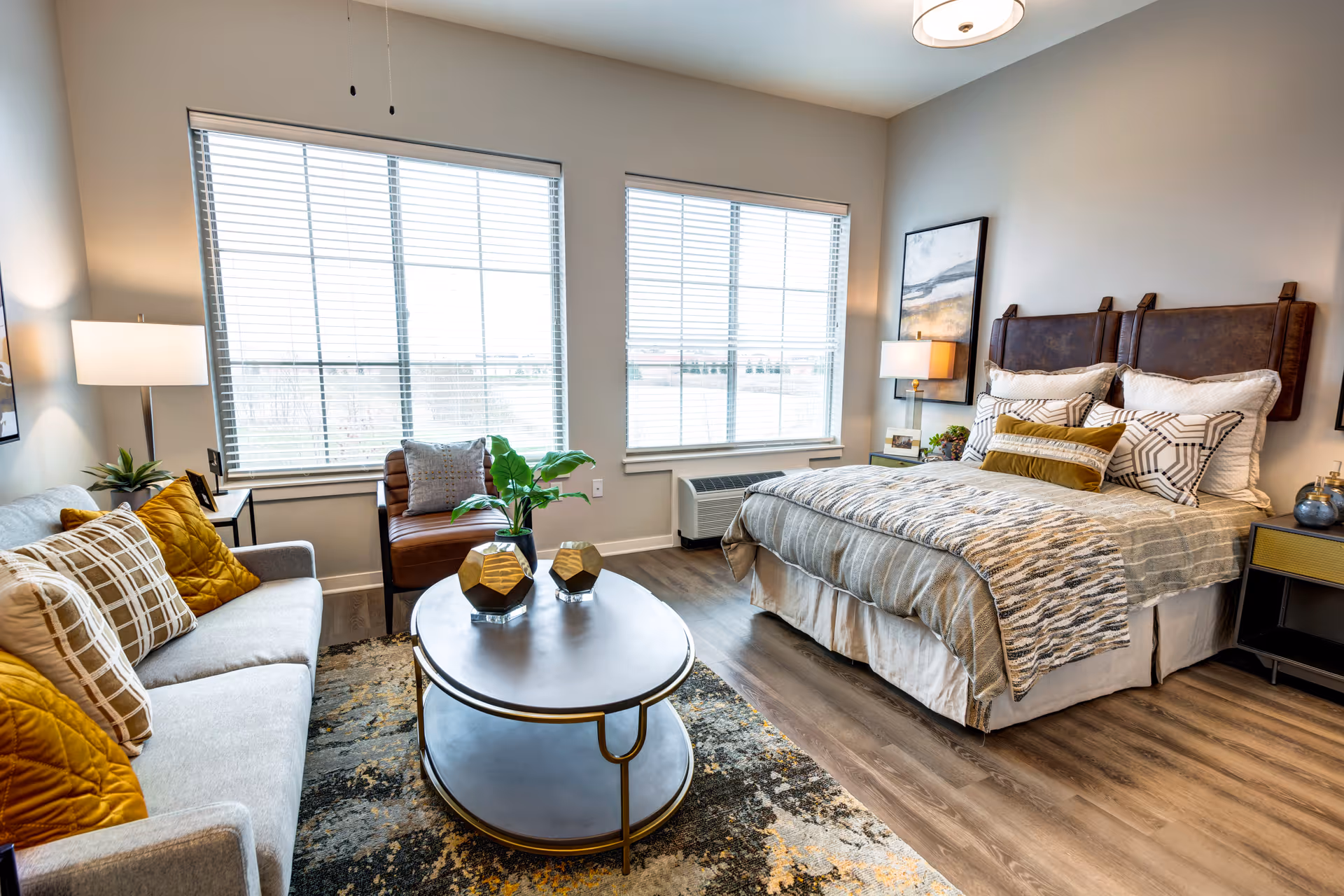 A bright, modern senior living bedroom featuring a large bed with multiple pillows and a patterned throw blanket. Next to the bed is a nightstand with a lamp and decorative items. Across from the bed is a gray sofa with mustard yellow and plaid pillows, a round coffee table with two gold geometric vases and a plant, and a brown leather chair near two large windows with white blinds. The room has wood flooring and neutral-colored walls with framed artwork.