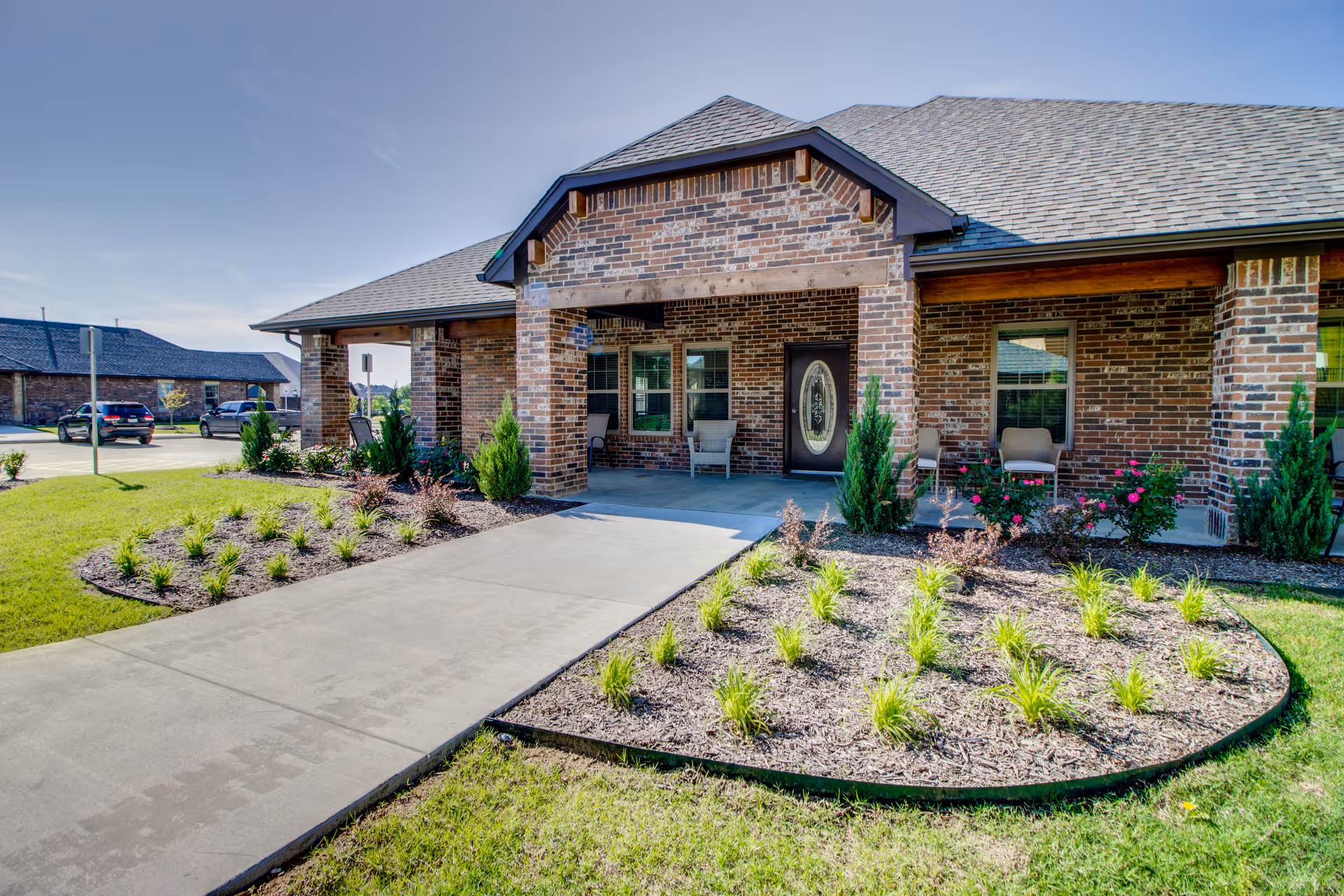 Front exterior view of a single-story brick building with a covered porch, chairs on the porch, landscaped garden beds with small plants and shrubs, and a concrete walkway leading to the entrance under a clear blue sky.