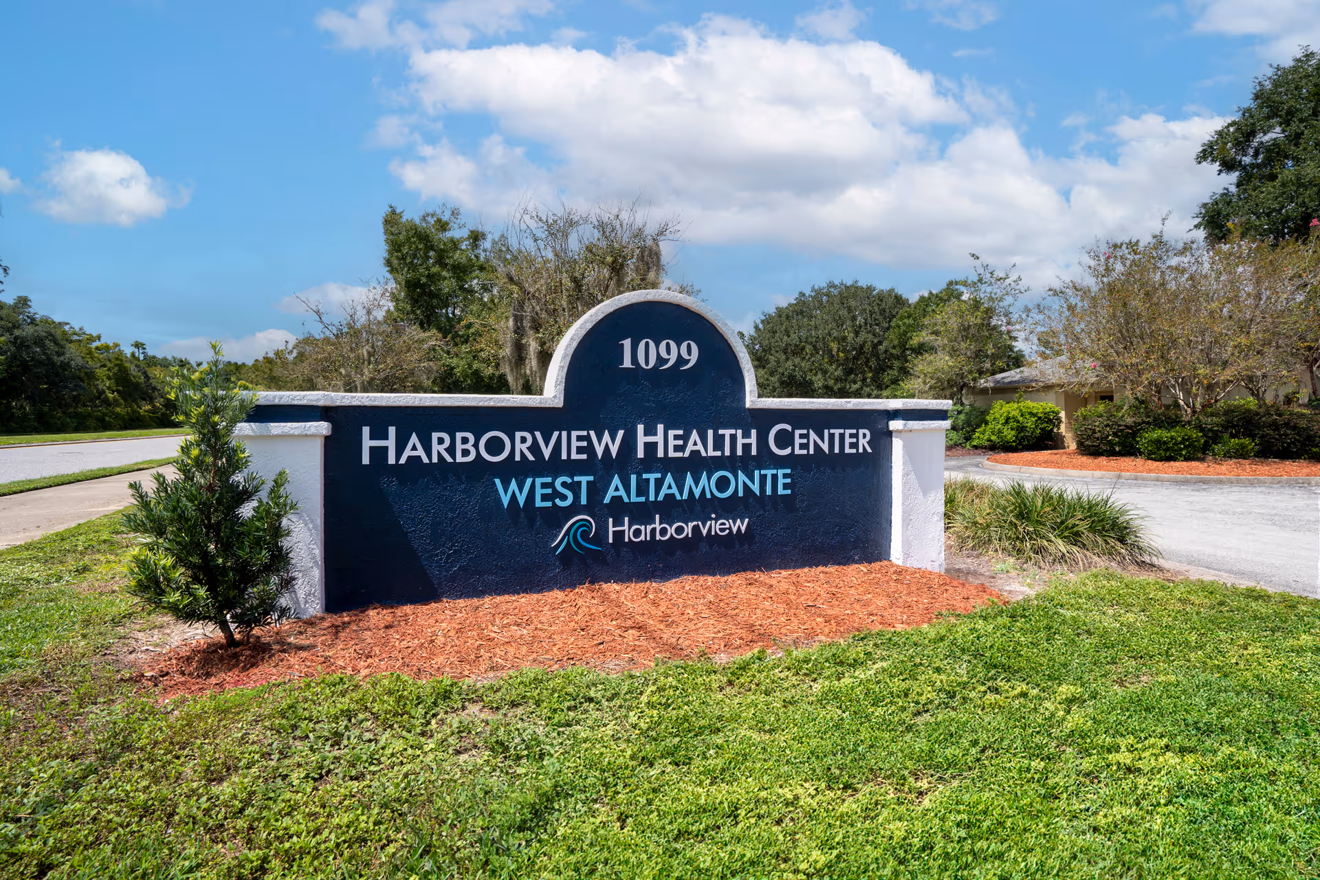 Stone entrance sign reading "Harborview Health Center West Altamonte" and the number 1099 set in landscaped grounds under a blue sky.