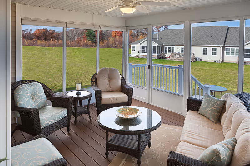 A cozy enclosed porch with wicker furniture including a cushioned sofa, two armchairs, and a glass-top coffee table. The porch has large windows offering a view of a grassy yard and neighboring buildings. The ceiling has a white fan with a light fixture.