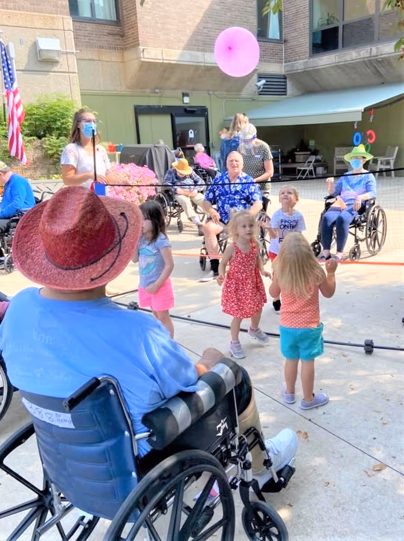 Seniors in wheelchairs and young children playing together outside a brick senior living facility courtyard during a daytime event.
