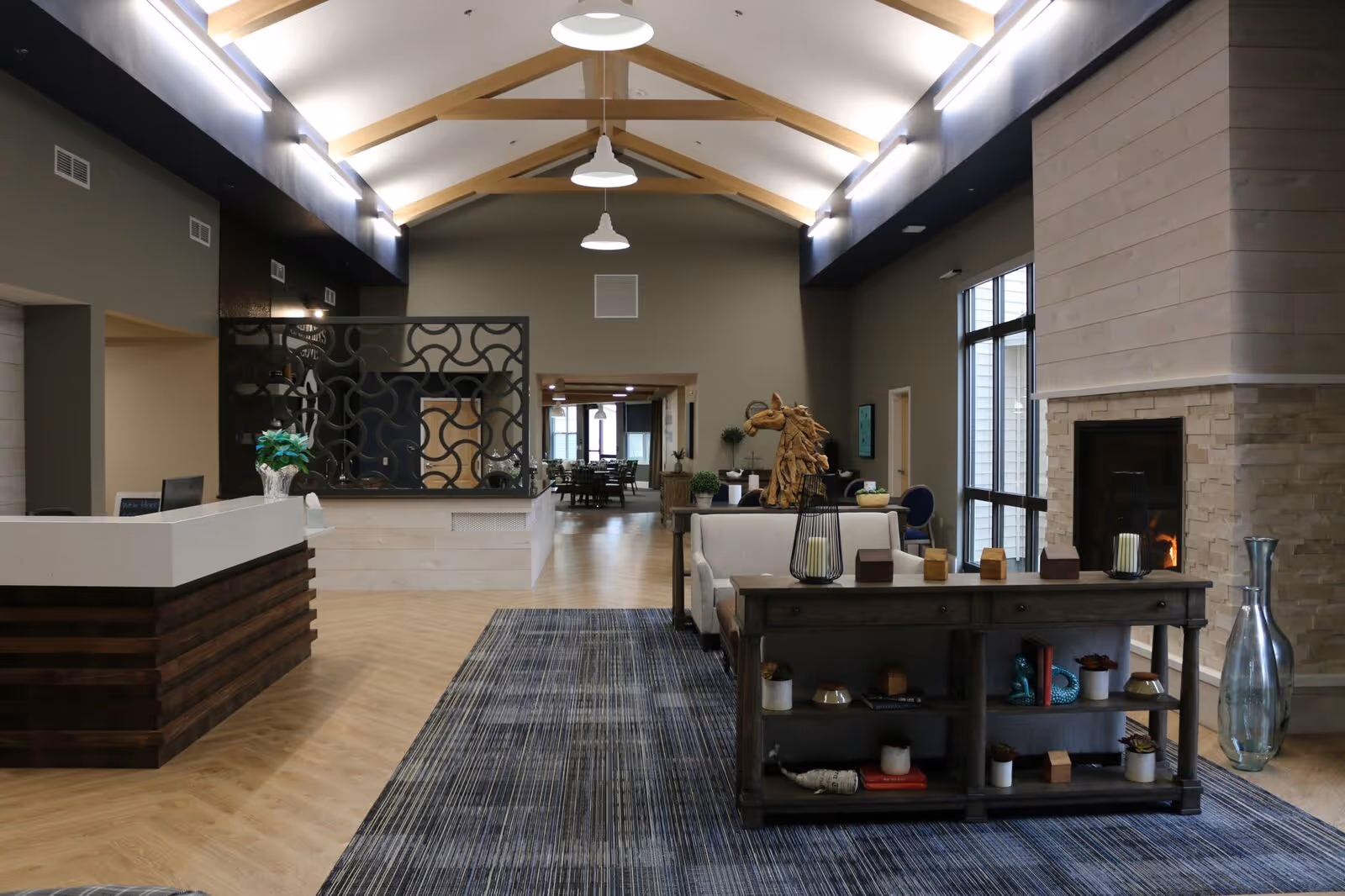 A spacious and modern senior living facility common area with high vaulted ceilings featuring wooden beams and pendant lights. The room includes a reception desk on the left, a decorative partition, a cozy seating area with a sofa and a wooden console table adorned with decorative items, and a fireplace on the right. Large windows allow natural light to enter, and a dining area is visible in the background.