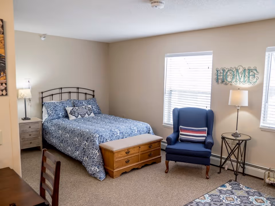 A cozy bedroom featuring a bed with blue patterned bedding, a wooden chest at the foot of the bed, a blue armchair with a striped pillow, a round side table with a lamp, two windows with white blinds, and a decorative wall sign that says 'HOME'.