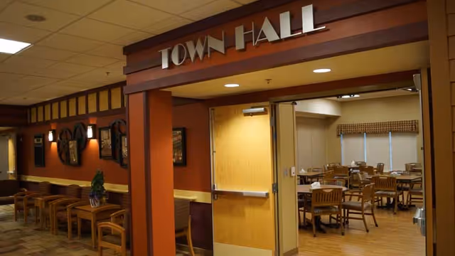 Interior view of a facility hallway leading to a room labeled 'Town Hall' with wooden chairs and tables inside. The hallway has wooden chairs lined up along the wall with framed decorations and warm lighting.