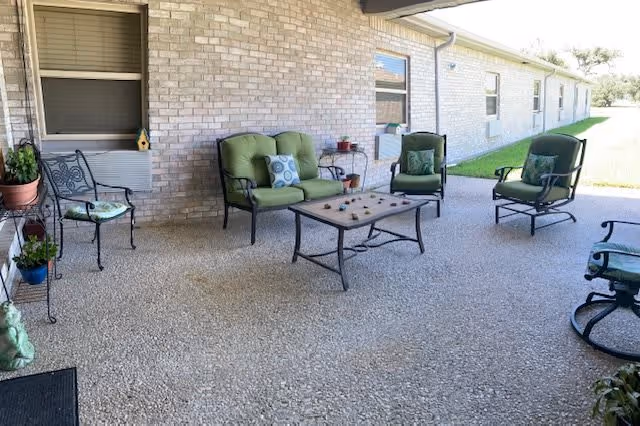 Outdoor patio area with a stone floor, featuring a set of green cushioned chairs and a loveseat around a rectangular coffee table. There are potted plants on metal stands and a brick wall with windows in the background.