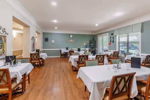 A dining room in an assisted living facility with several tables covered in white tablecloths, each set with glasses and utensils. The room has wooden floors, green walls with white trim, and large windows with blinds allowing natural light to enter. Chairs with wooden frames and cushioned seats surround the tables. There are decorative items on the walls and a clock above a sign on the far wall.