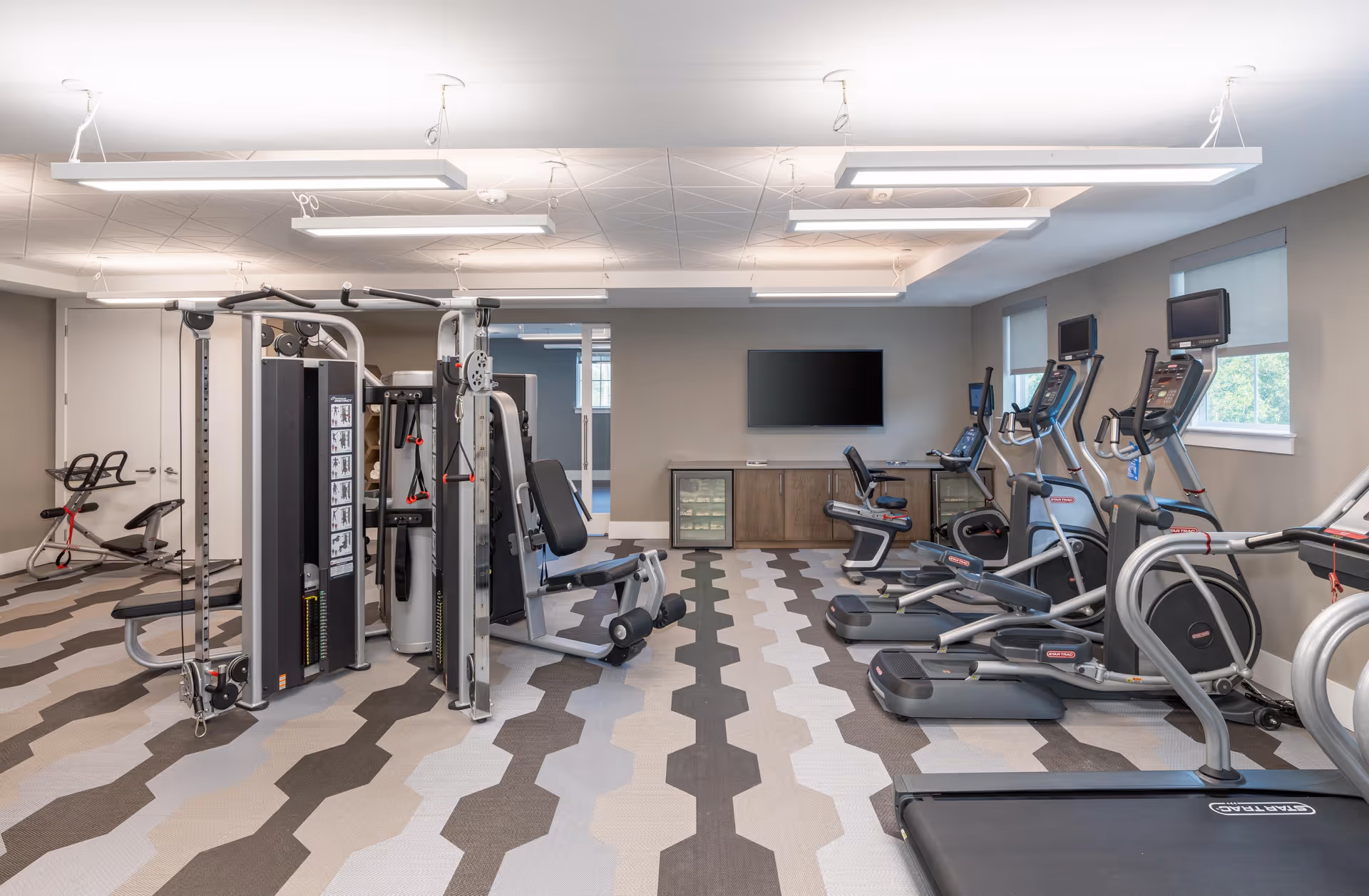 A modern fitness room with various exercise equipment including treadmills, elliptical machines, stationary bikes, and a multi-functional weight machine. The room has a patterned floor with shades of gray and beige, large windows with blinds, a wall-mounted flat screen TV, and overhead rectangular light fixtures.