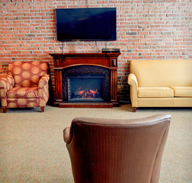 A cozy living room with a brick wall background featuring a mounted flat-screen TV above a wooden electric fireplace. There is a patterned armchair on the left, a beige sofa on the right, and a brown leather chair in the foreground.