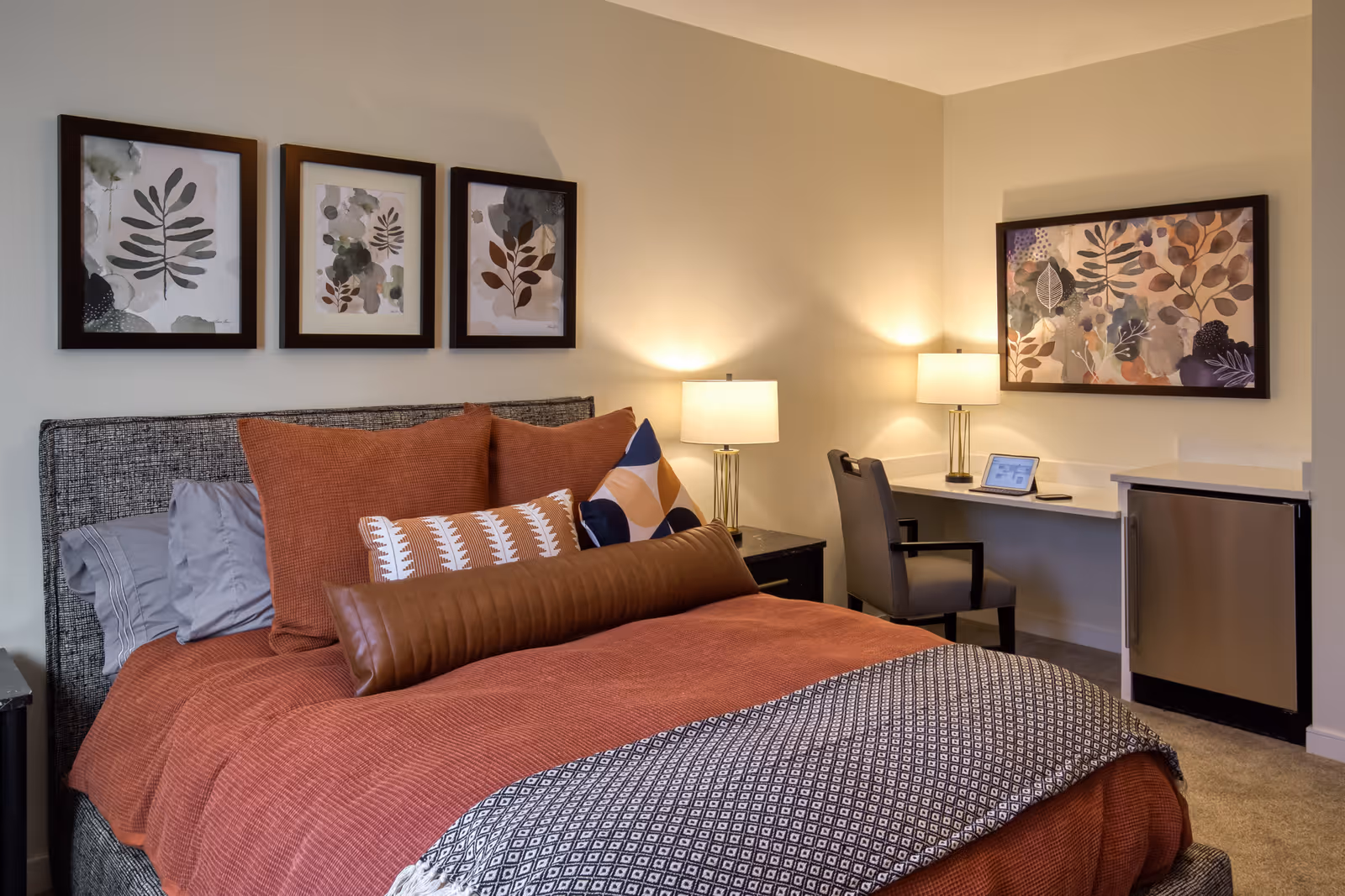 Cozy furnished bedroom with a made bed featuring patterned pillows, two bedside lamps, framed botanical artwork, a desk with a chair and a mini-fridge.