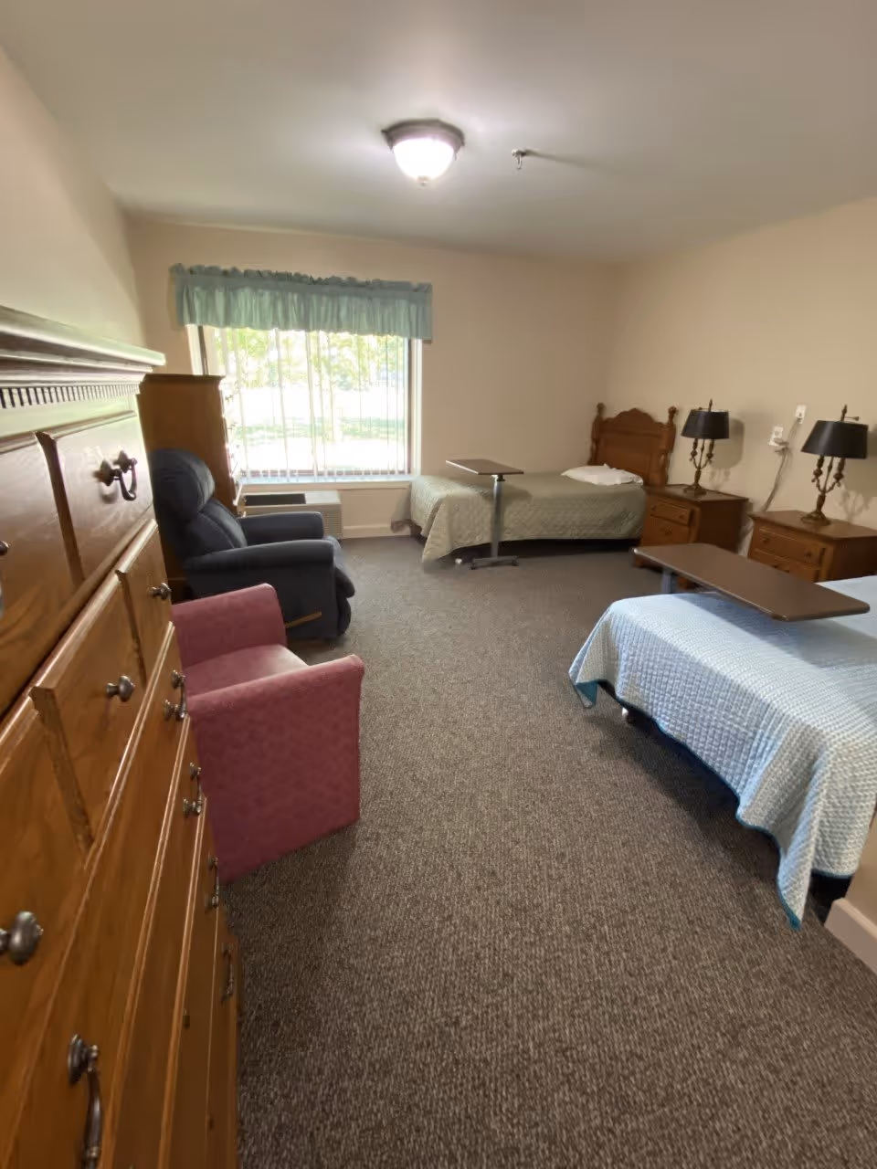 A senior living facility bedroom with two single beds, each with a small overbed table. The room has a large window with vertical blinds and a light blue valance. There are two wooden nightstands with matching lamps beside one bed, a wooden dresser on the left side, and two armchairs, one dark blue and one pink, near the window. The floor is carpeted and the walls are painted a light beige color.