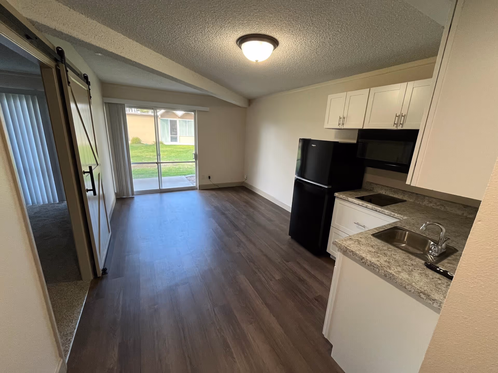 Interior view of a small apartment unit with a kitchenette on the right featuring white cabinets, a black refrigerator, a microwave, a small sink, and a cooktop. The room has wood flooring and a sliding glass door at the far end leading to a grassy outdoor area. To the left, there is a doorway with a sliding barn door that opens to a carpeted bedroom.