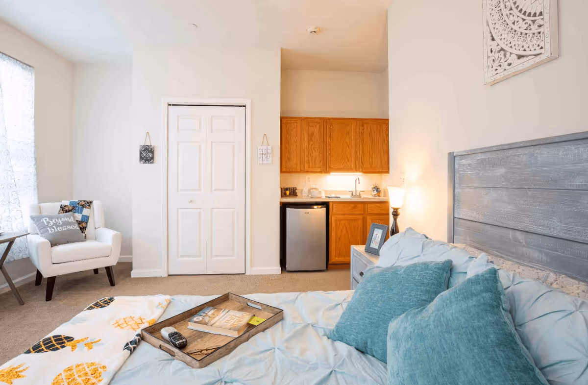A cozy senior living apartment bedroom with a bed featuring blue pillows and a light blue comforter. A wooden tray with a book, glasses, and a remote control rests on the bed. To the right, there is a nightstand with a lamp and a framed photo. In the background, there is a small kitchenette with wooden cabinets, a mini fridge, and a sink. To the left, a white armchair with a pillow that says 'Beyond blessed' sits next to a window with sheer curtains. The walls are painted light beige, and there is a decorative wall hanging above the bed.