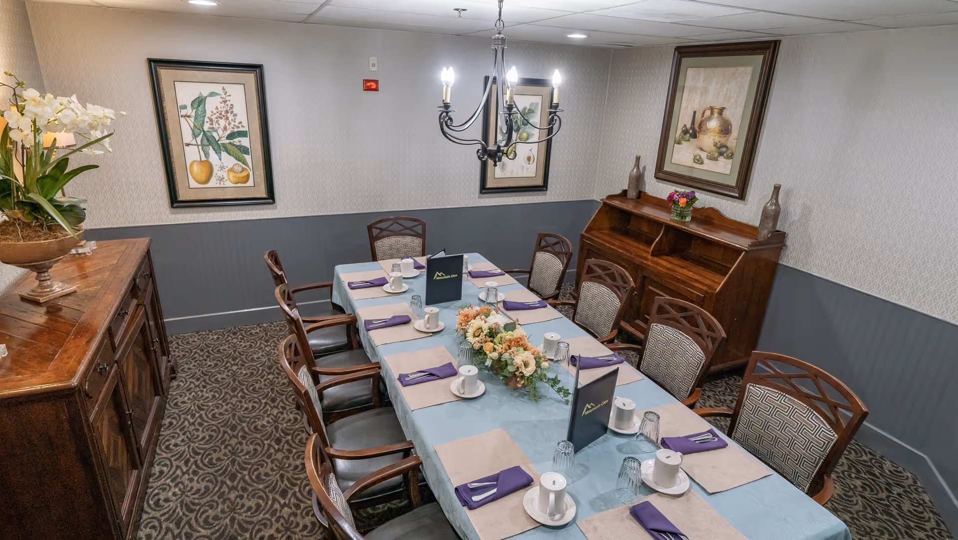 A dining room with a long table set for eight people. The table is covered with a light blue tablecloth and has beige placemats, white cups and saucers, upside-down glasses, and purple napkins. A floral centerpiece decorates the middle of the table. The room has patterned carpet, light gray walls with a darker gray wainscoting, and framed botanical artwork on the walls. There is a wooden sideboard with a flower arrangement on the left and a wooden cabinet with decorative items on the right. A chandelier with lit bulbs hangs above the table.