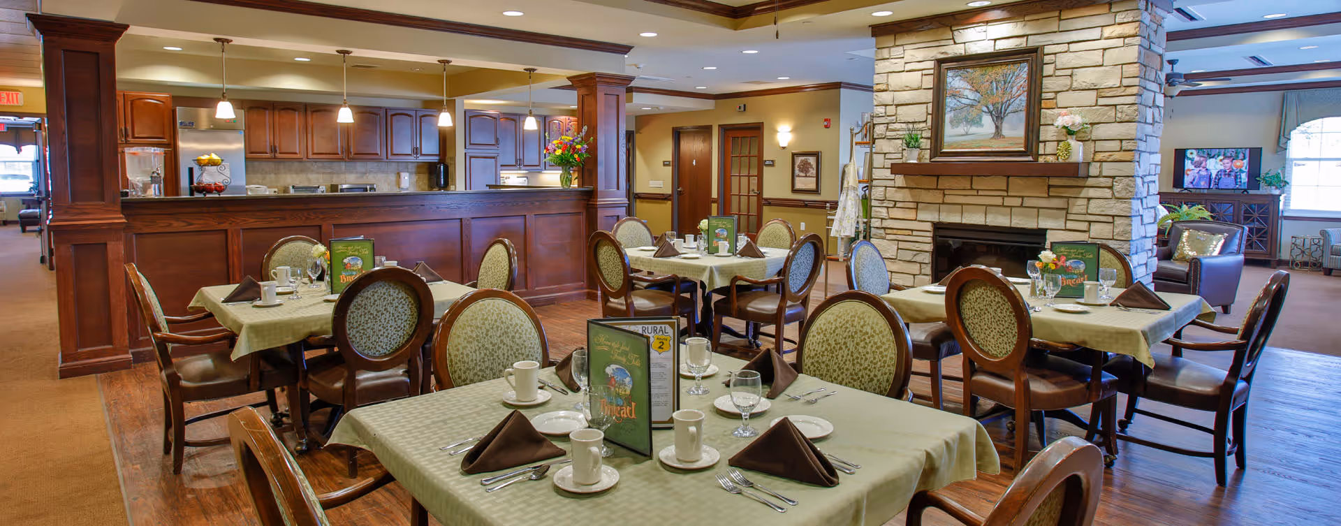A dining room in a senior living facility with several tables covered with light green tablecloths, each set with cups, glasses, silverware, and folded brown napkins. The room features wooden chairs with patterned upholstery, a stone fireplace with a painting above it, and a wooden counter area in the background. The space is warmly lit with ceiling lights and has a cozy, inviting atmosphere.