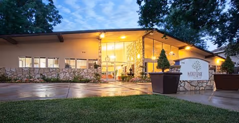 Front entrance of McKinley Park Care Center at dusk with a lit canopy, stone facade, planters, and a lawn.
