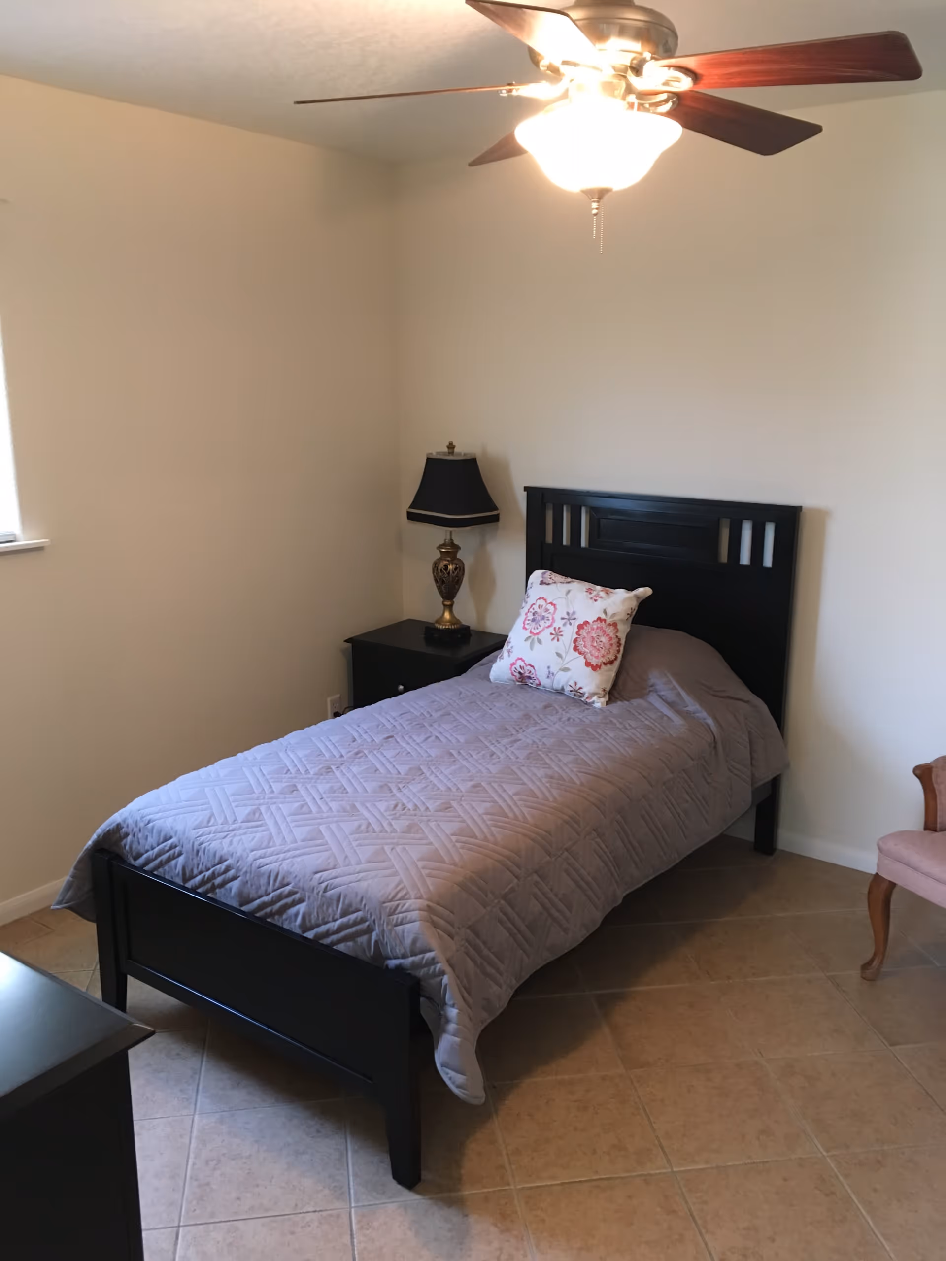 A simple bedroom with a single black bed frame, a gray quilted bedspread, and a floral pillow. There is a black nightstand with a decorative lamp beside the bed, a ceiling fan with lights above, and a pink upholstered chair partially visible on the right. The floor is tiled and the walls are plain light-colored.