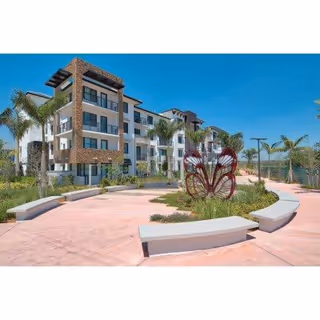 Modern multi-story residential building with balconies, surrounded by palm trees and landscaped garden featuring a large butterfly sculpture. The sky is clear and blue.