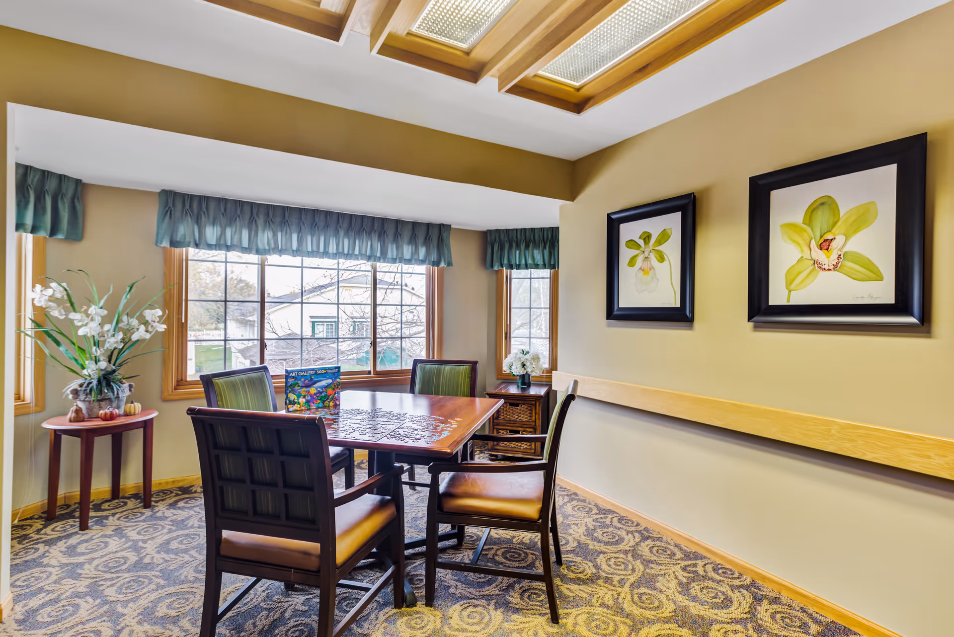 Small dining area with a wooden table and chairs under skylights, floral artwork on the wall, and windows with green valances.
