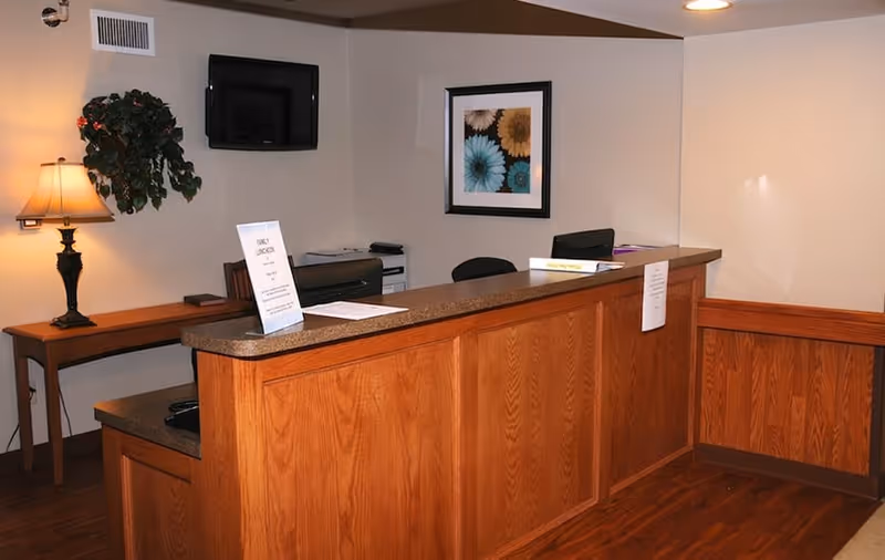Reception area with a wooden front desk, a table lamp on a side table, a wall-mounted TV, a framed floral picture on the wall, and some papers and signs on the desk.