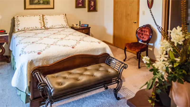 A cozy bedroom featuring a wooden bed with a white quilted bedspread decorated with floral patterns. At the foot of the bed is a cushioned bench with a tufted leather seat and ornate metal armrests. To the right, there is a vintage-style red velvet chair and a wooden door. A vase with white flowers and greenery is visible on the right side, and framed pictures hang on the wall above the bed.