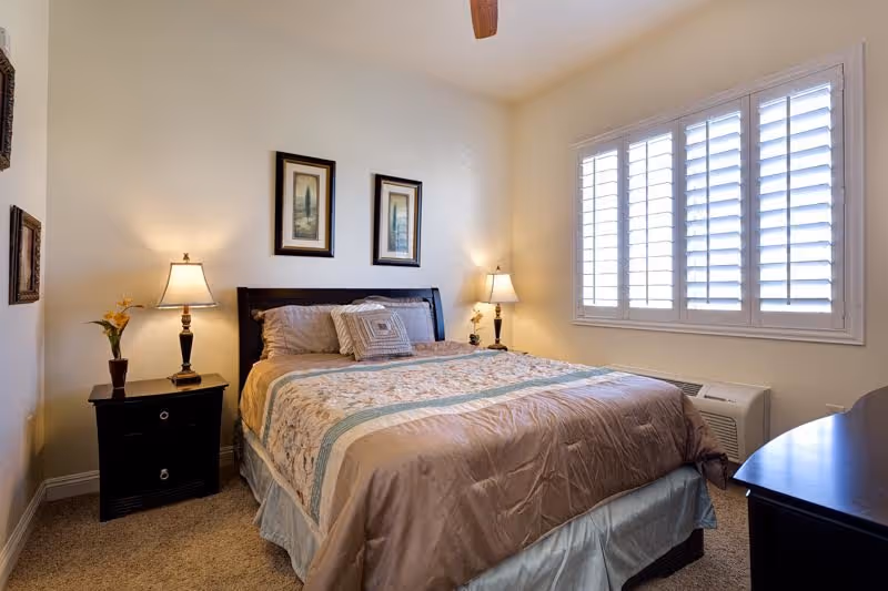 Well-lit bedroom with a double bed, matching nightstands and lamps, framed artwork, and a window with shutters.