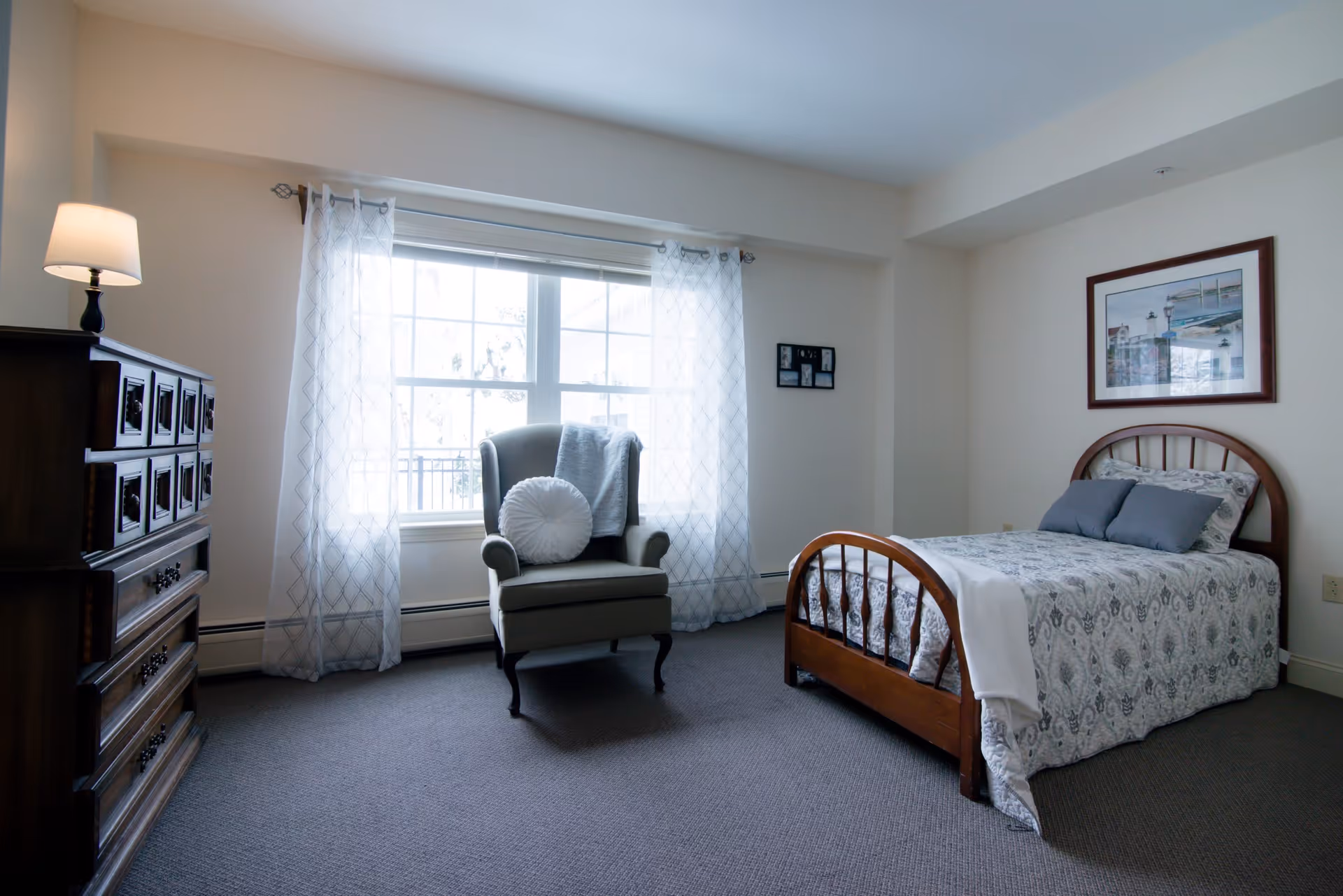 A bright bedroom with a single wooden bed covered in patterned bedding and two pillows. Next to the bed is a framed picture on the wall. A cushioned armchair with a round pillow and a blanket is placed near a large window with sheer white curtains. A wooden dresser with a lamp on top is positioned on the left side of the room.