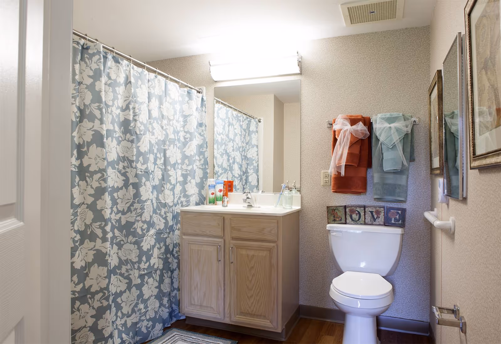 A clean bathroom featuring a white toilet with decorative blocks spelling 'LOVE' on the tank, a vanity with a sink and toiletries, a large mirror above the sink, a blue and white floral shower curtain, and two sets of towels hanging on the wall, one orange and one light blue, each tied with a white ribbon.