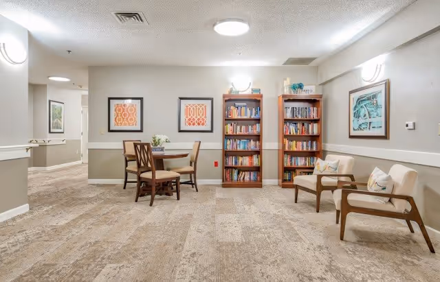 A cozy interior common area with two wooden bookshelves filled with books, a round wooden table with four chairs, and two cushioned armchairs with decorative pillows. The walls are adorned with framed artwork, and the space is well-lit with ceiling and wall lights. The floor is carpeted with a neutral pattern.