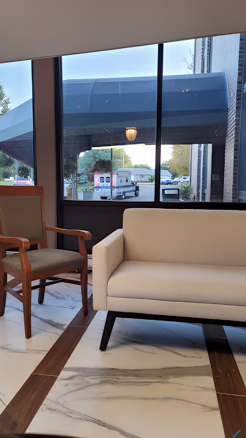 Interior view of a waiting area with a beige sofa and a wooden armchair with green upholstery, large windows showing an ambulance outside under a covered entrance.