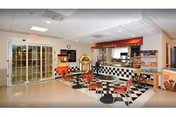 Interior view of a retro-style dining area with black and white checkered flooring and red chairs. There is a counter with a red awning labeled 'SNACKS' and a jukebox near the entrance with glass doors. The space is well-lit with ceiling lights and has a clean, polished floor.